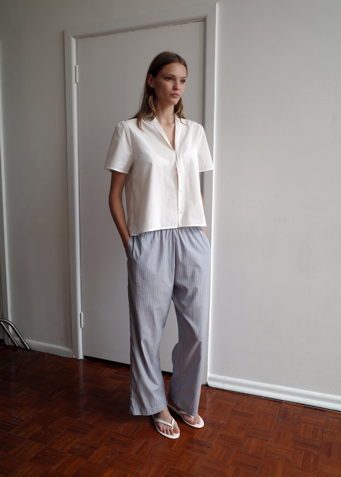 A woman stands indoors on a wooden floor against a white wall, hands in pockets, wearing a short-sleeved white button-up shirt, white sandals, and the Ease Trouser - Glacier Stripe from Deiji Studios.