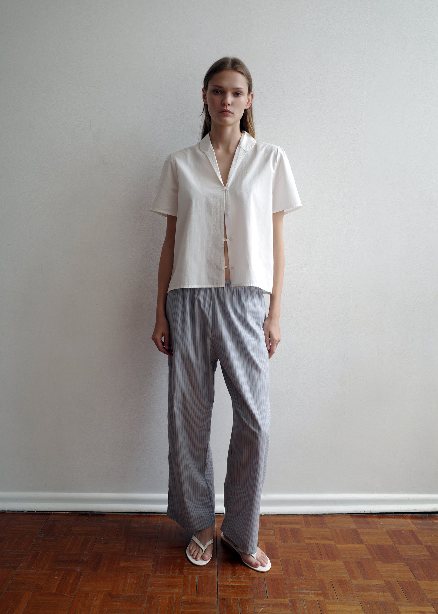 A person stands indoors against a plain white wall, wearing a short-sleeved white blouse, Deiji Studios Ease Trouser - Glacier Stripe, and white sandals on a brown wooden floor.
