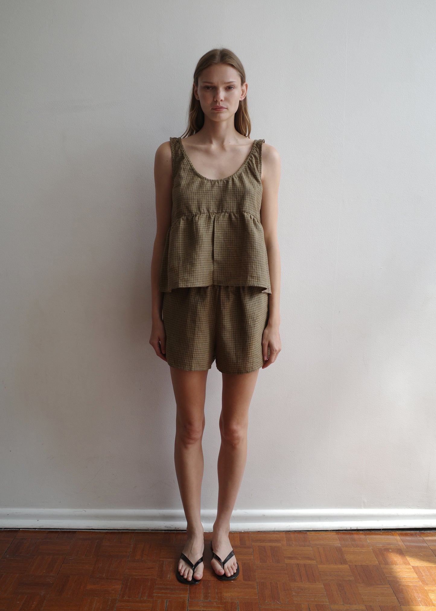 A woman stands indoors against a white wall, wearing the Gathered Linen Set - Pine Check from Deiji Studios with black flip-flops. She has straight hair and a neutral expression.