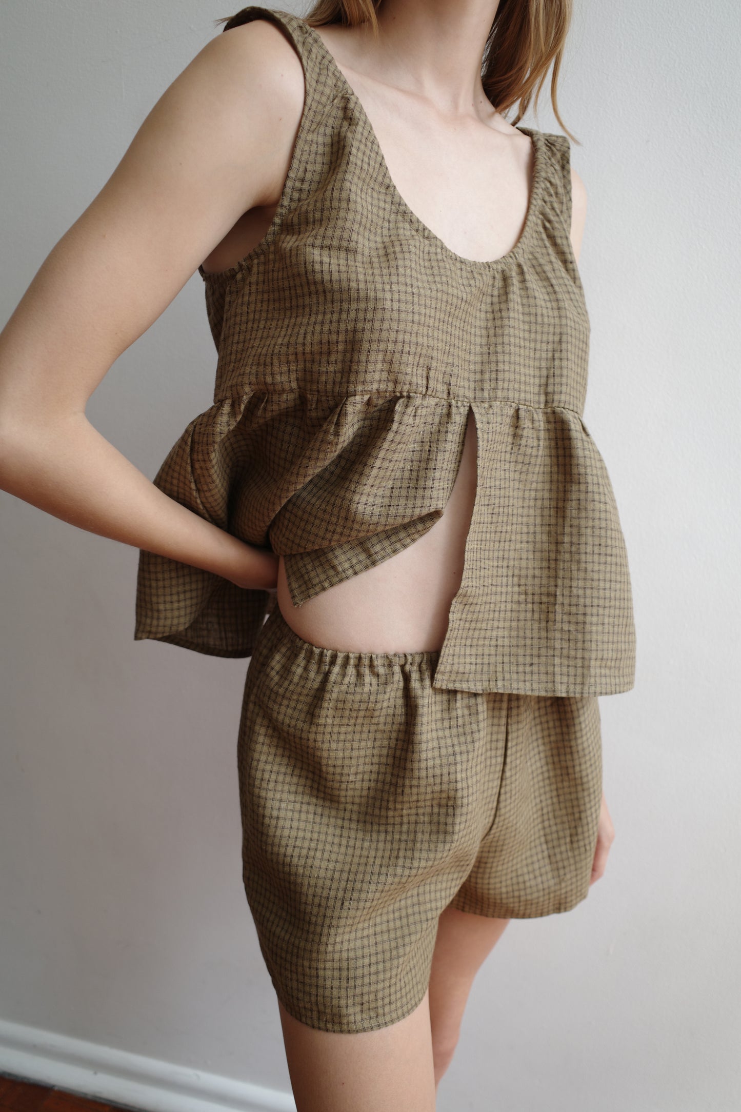 A person wearing Deiji Studios’ Gathered Linen Set in Pine Check—a brown, sleeveless, loose-fitting top with a front slit and matching high-waisted shorts—stands against a plain white wall.
