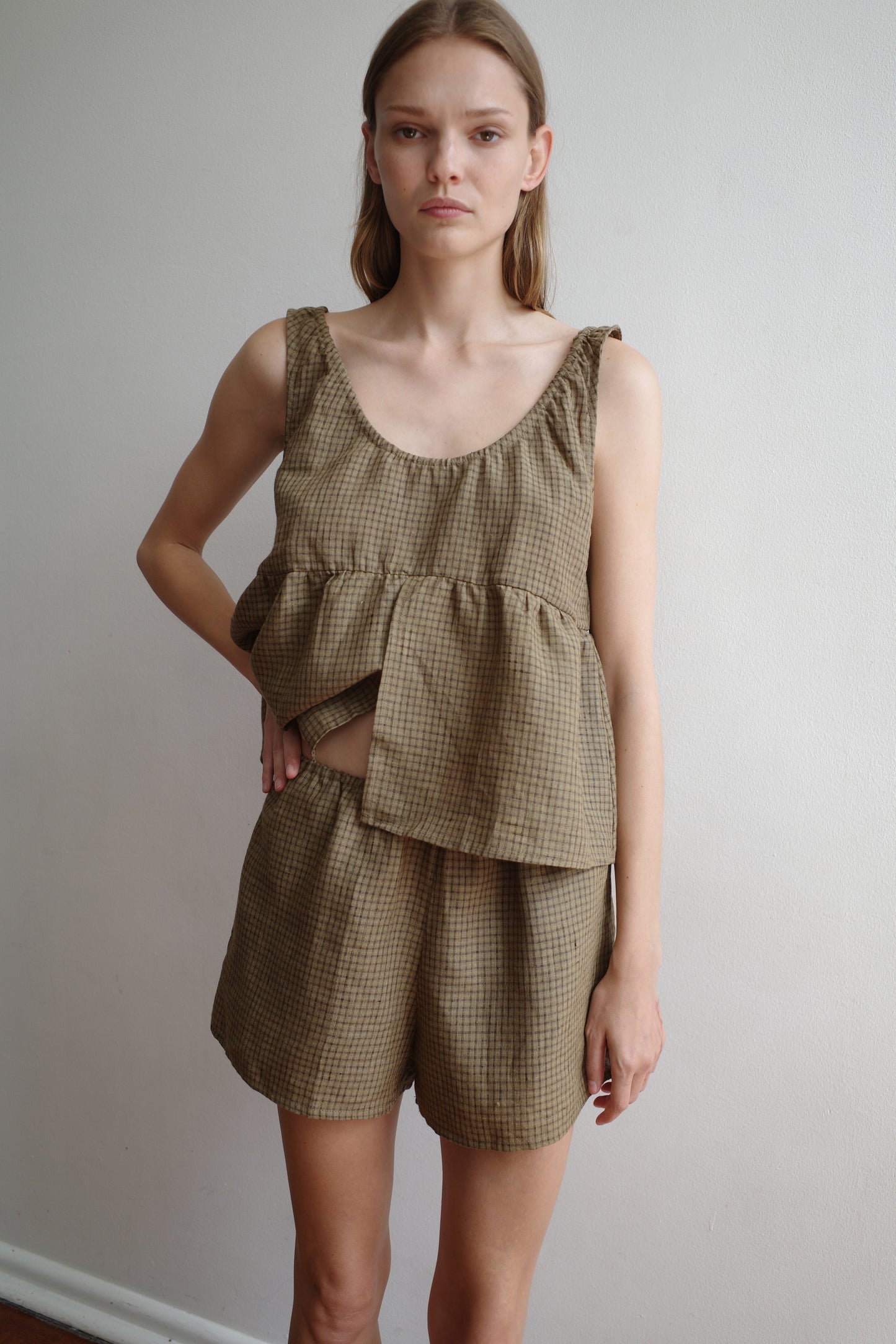 A woman with straight light brown hair and a neutral expression wears the Deiji Studios Gathered Linen Set in Pine Check—a loose, sleeveless brown plaid linen outfit with ruffle detail—against a plain, light-colored wall.