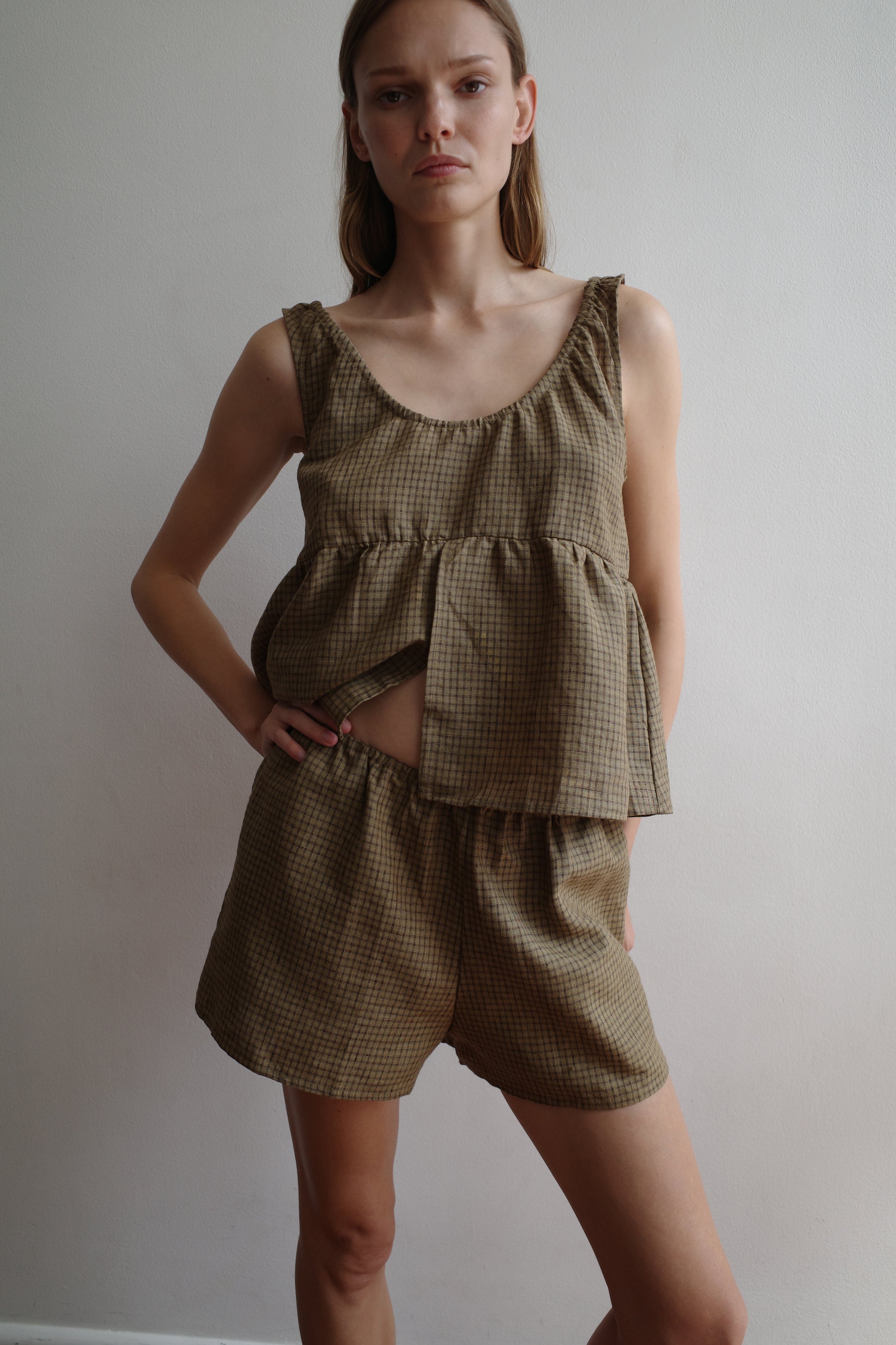 A woman stands against a plain white wall, wearing Deiji Studios’ Gathered Linen Set in Pine Check. The loose, sleeveless plaid top and matching shorts give a relaxed look. She faces the camera neutrally with one hand on her hip.