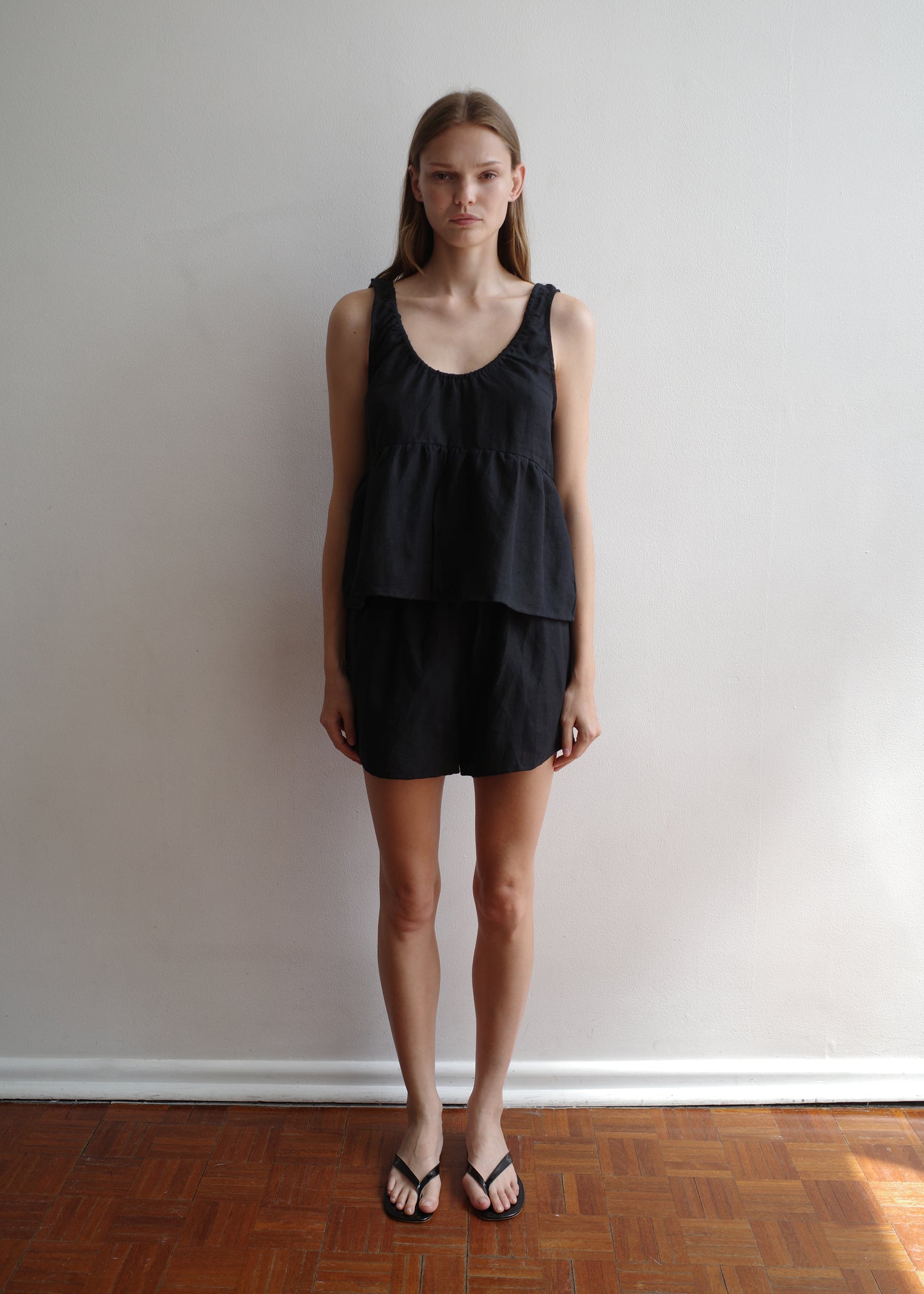 A woman stands indoors against a white wall and wooden floor, wearing the Gathered Linen Set - Black from Deiji Studios—a sleeveless layered top with matching shorts—and black flip-flops, looking straight at the camera.