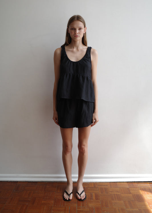 A woman stands indoors against a white wall and wooden floor, wearing the Gathered Linen Set - Black from Deiji Studios—a sleeveless layered top with matching shorts—and black flip-flops, looking straight at the camera.