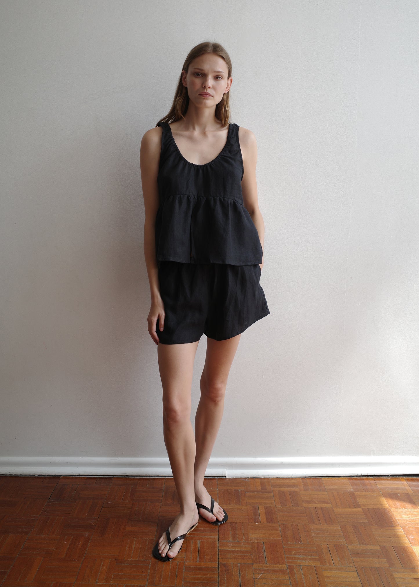 A person stands indoors against a white wall, wearing the Gathered Linen Set - Black by Deiji Studios with black flip-flops on a wooden floor. They have straight light brown hair and a neutral expression.