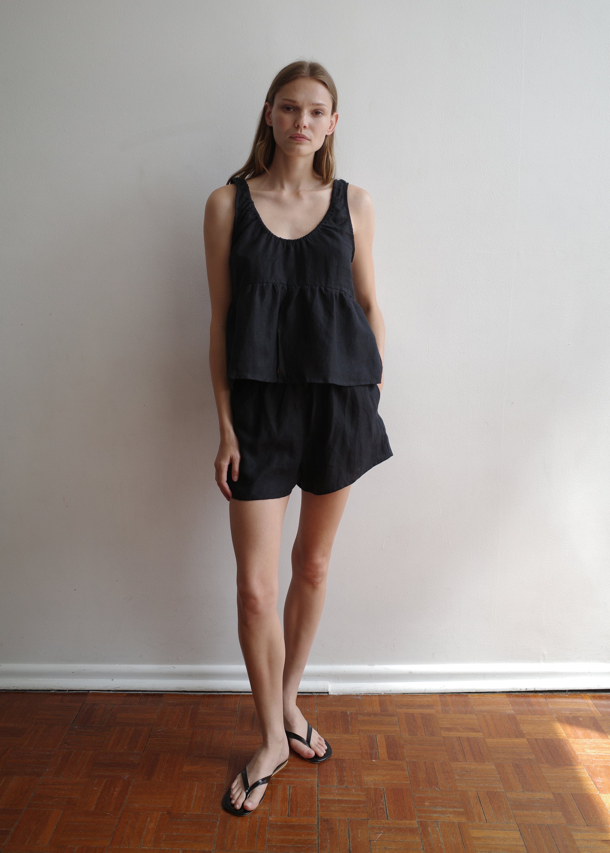 A person stands indoors against a white wall, wearing the Gathered Linen Set - Black by Deiji Studios with black flip-flops on a wooden floor. They have straight light brown hair and a neutral expression.