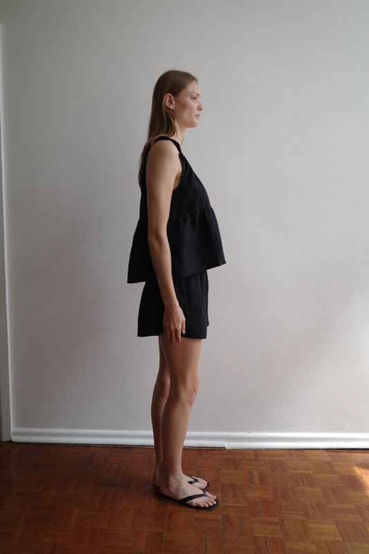 A person with long hair stands in profile against a plain white wall, wearing Deiji Studios' Gathered Linen Set - Black, paired with black sandals on a wooden floor.