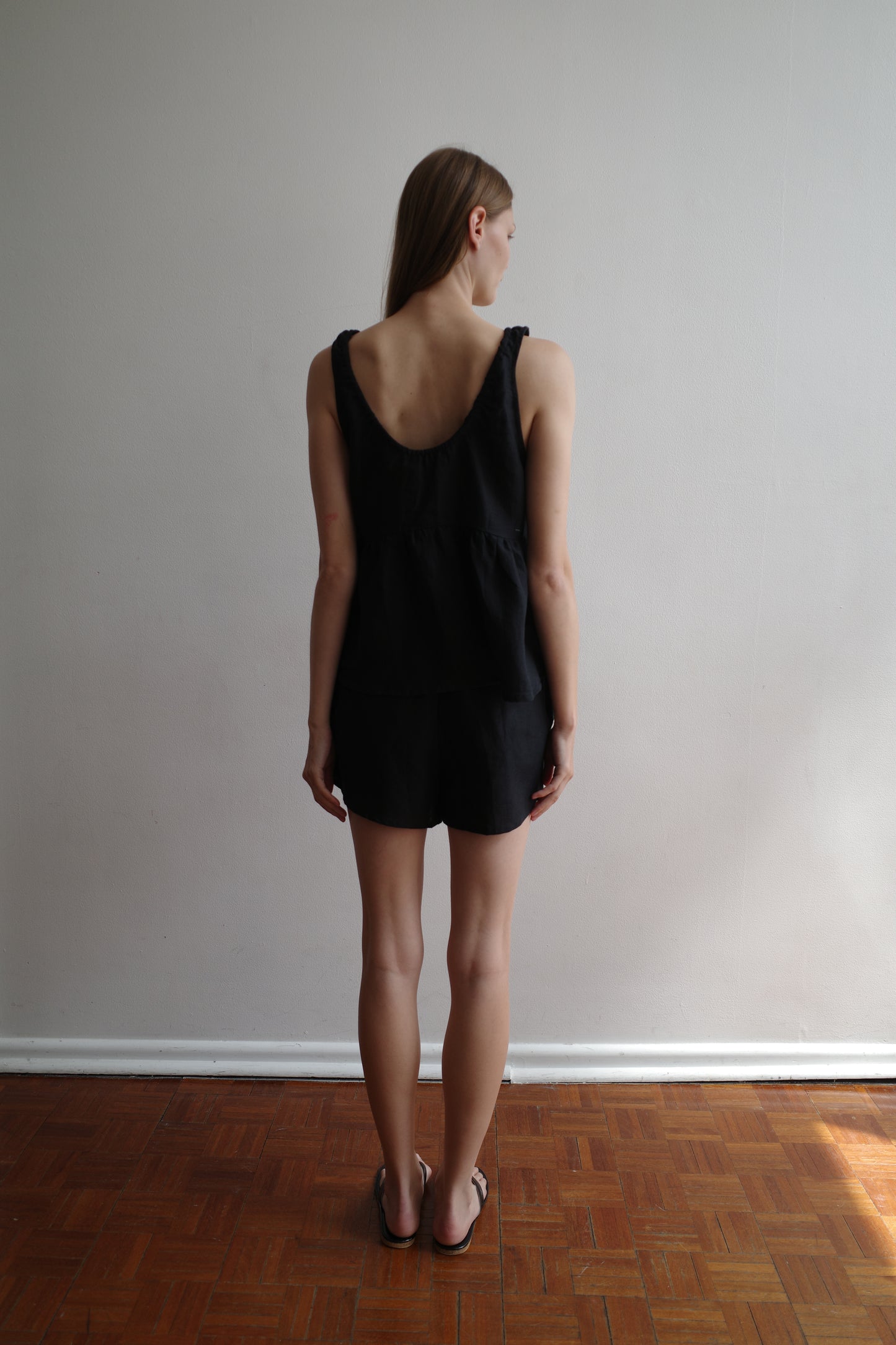 A woman stands with her back to the camera, wearing the Gathered Linen Set - Black by Deiji Studios with sandals, against a plain light wall and wooden floor.