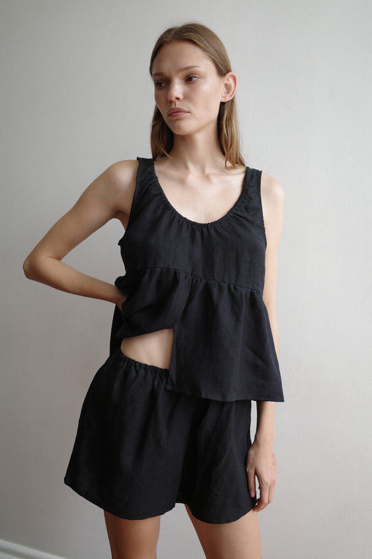 A woman with straight light brown hair and a neutral expression stands against a plain wall, wearing the Gathered Linen Set - Black by Deiji Studios, featuring a loose sleeveless top with an asymmetrical hem and matching shorts in organic French linen.