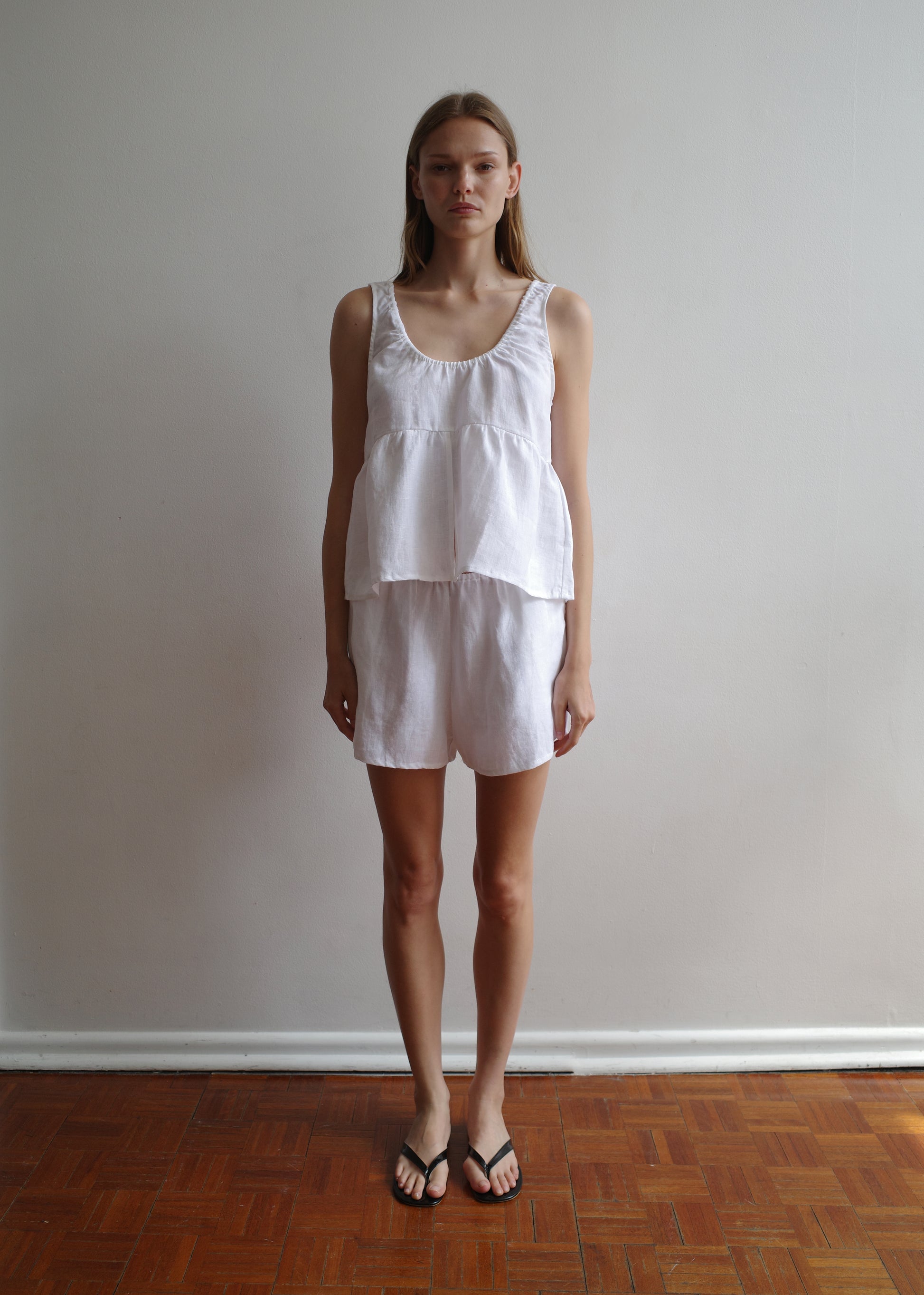 A woman with straight hair and a neutral expression stands indoors against a plain light wall, wearing the Gathered Linen Set - White by Deiji Studios and black flip-flops, on a wooden floor.