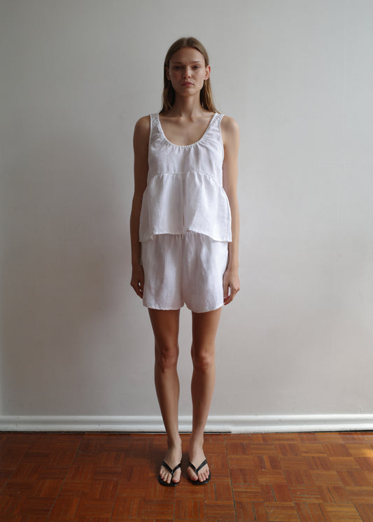 A woman with straight hair and a neutral expression stands indoors against a plain light wall, wearing the Gathered Linen Set - White by Deiji Studios and black flip-flops, on a wooden floor.