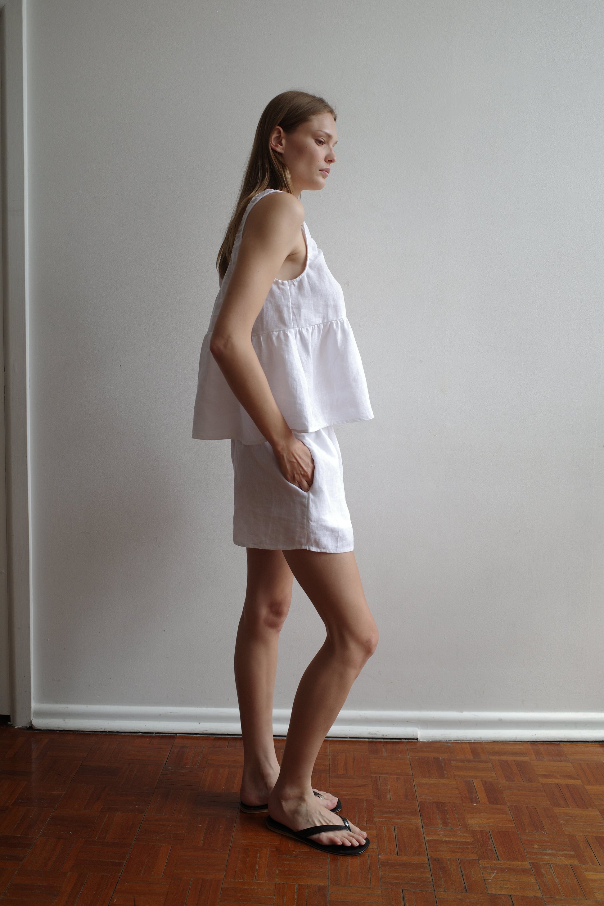 A woman stands sideways against a plain white wall, wearing the Gathered Linen Set - White by Deiji Studios in organic French linen. Her long hair is straight, left hand in her pocket, black sandals on her feet, and she stands on a wooden floor.