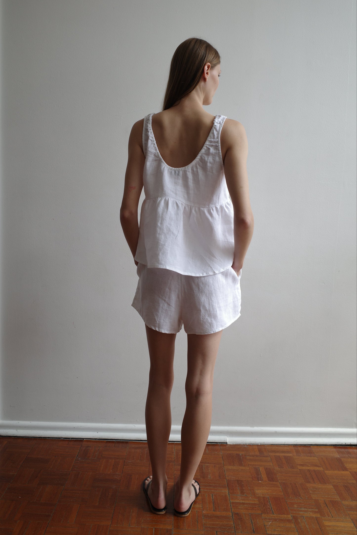 A woman with long hair, seen from behind, wears the Gathered Linen Set - White by Deiji Studios. The sleeveless top and shorts have pockets. She stands on a wooden floor against a plain light-colored wall.