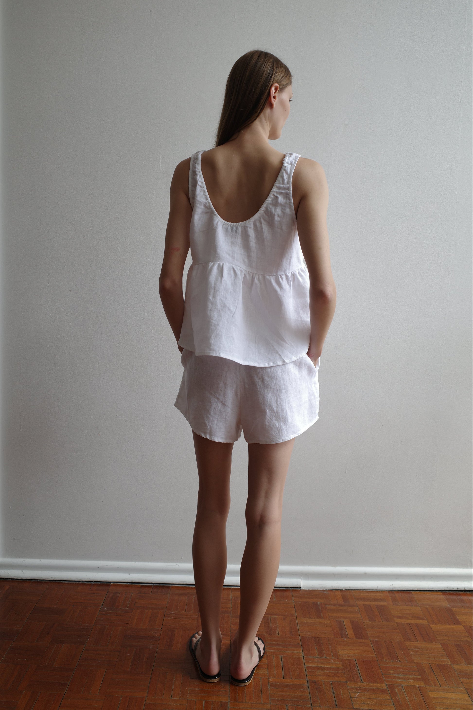 A woman with long hair, seen from behind, wears the Gathered Linen Set - White by Deiji Studios. The sleeveless top and shorts have pockets. She stands on a wooden floor against a plain light-colored wall.
