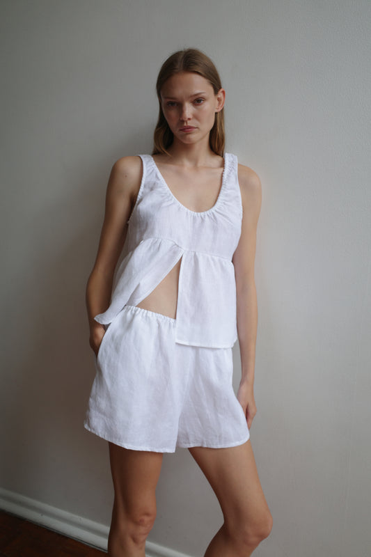 A woman stands against a plain wall, wearing the Gathered Linen Set - White by Deiji Studios—sleeveless, with an asymmetric cutout and matching shorts made from organic French linen—looking directly at the camera with a neutral expression.