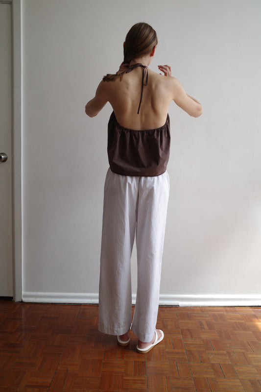A person with long hair faces a white wall, wearing a brown halter backless top, Ease Trouser Sage Stripe by Deiji Studios in loose white, and white shoes on a wooden floor.