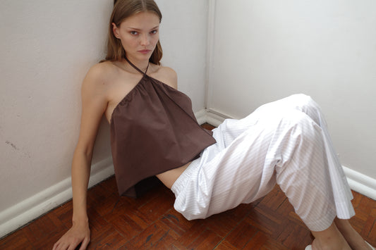 A young woman with straight light brown hair sits on a wooden floor in a corner, wearing a brown halter top and Ease Trouser Sage Stripe by Deiji Studios, gazing seriously at the camera against plain white walls.