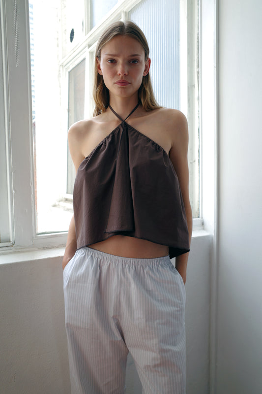 A woman stands indoors by a window, wearing a dark halter top and the Ease Trouser Sage Stripe from Deiji Studios—light, loose-fitting organic cotton pants with her hands in her pockets as natural light filters through behind her.