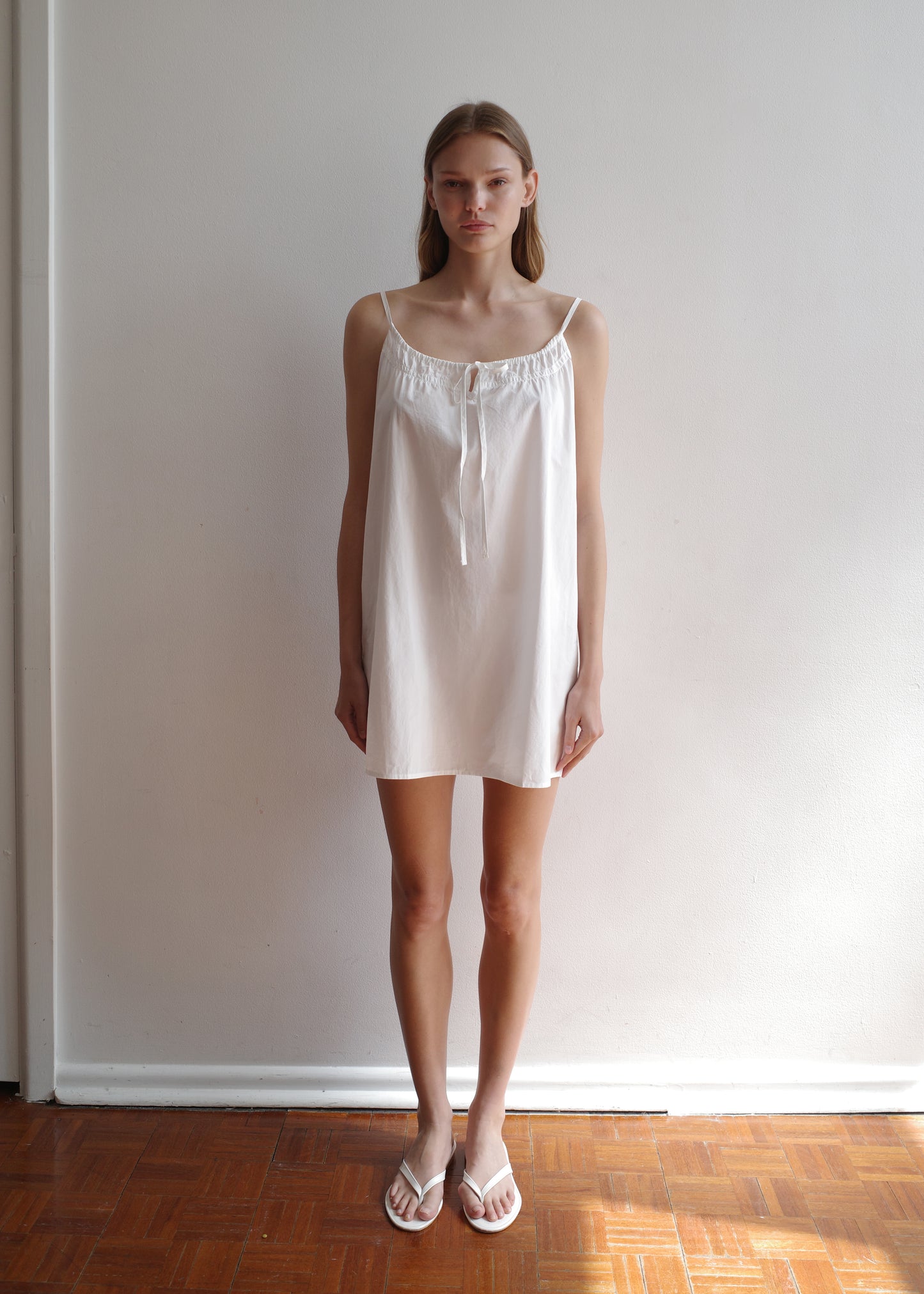 A woman stands indoors against a plain white wall, wearing the Light Dress - White, a light mini dress with thin straps and a front tie, paired with white sandals. Sunlight highlights the wooden floor near her feet.