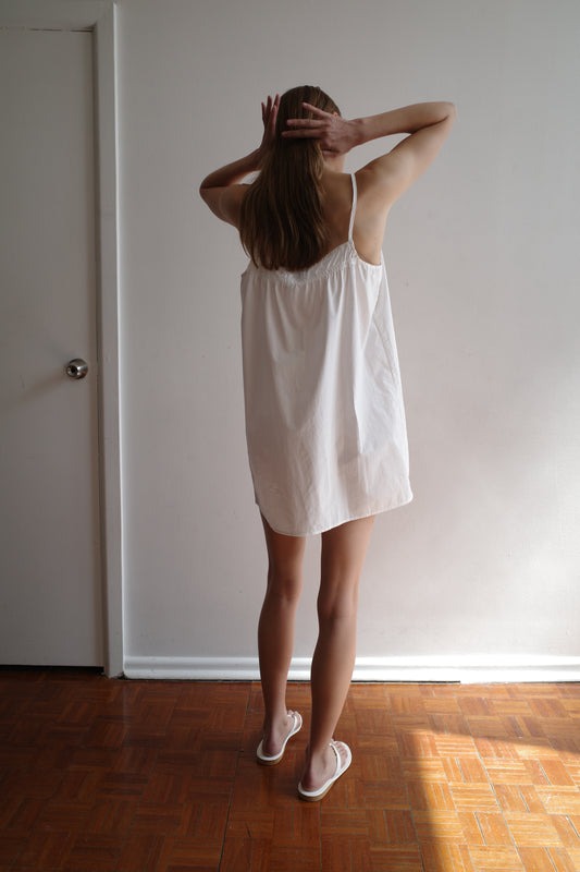 A person wearing the Light Dress - White and sandals stands on a wooden floor facing a white wall, hands behind their head, with their back to the camera.