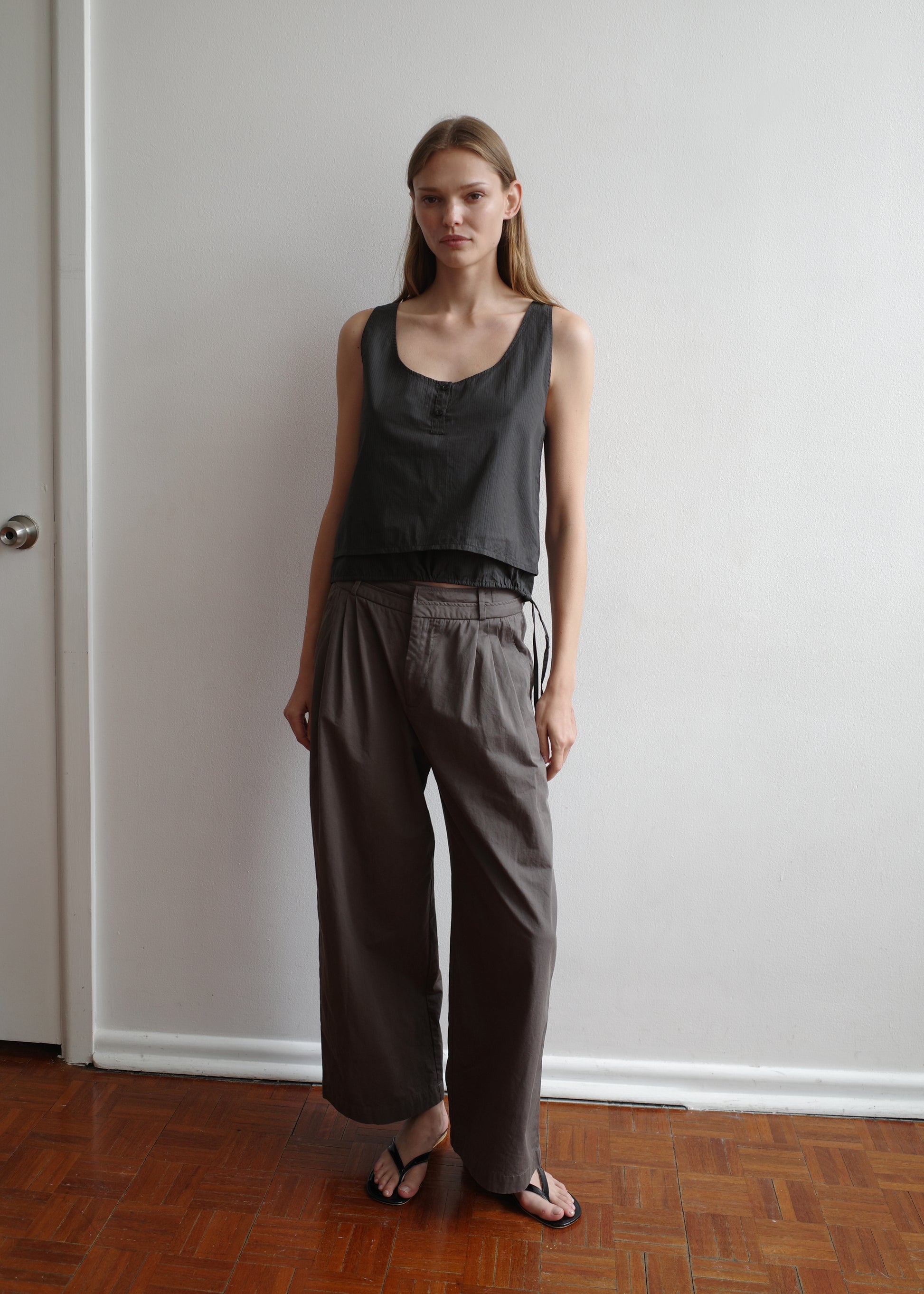 A woman stands indoors by a white wall, wearing a sleeveless Deiji Studios Layered Top - Black Stripe, loose gray belted pants, and black sandals. She has straight light brown hair and a neutral expression.