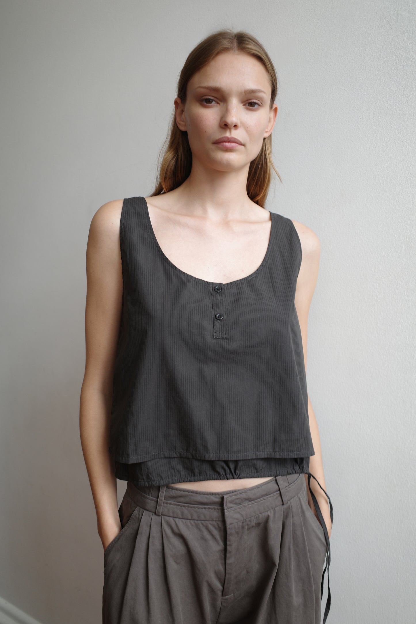 A woman stands against a plain light wall, wearing Deiji Studios’ sleeveless Layered Top in Black Stripe and loose, high-waisted taupe pants. She has light brown hair, a neutral expression, and her hands in her pockets.