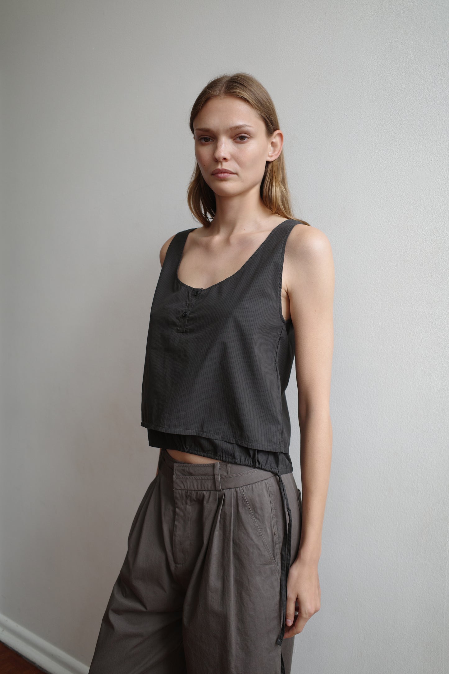 A woman with straight blond hair wears the Layered Top - Black Stripe, a sleeveless organic cotton piece, and loose gray pants, standing against a plain white wall and looking calmly at the camera.