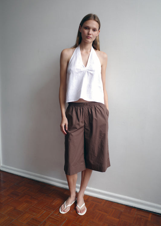 A woman stands indoors against a plain wall, wearing a white halter top, white sandals, and the Long Lounger - Carob Check: loose, brown checked organic cotton knee-length shorts with pockets. She has straight light brown hair and a neutral expression.