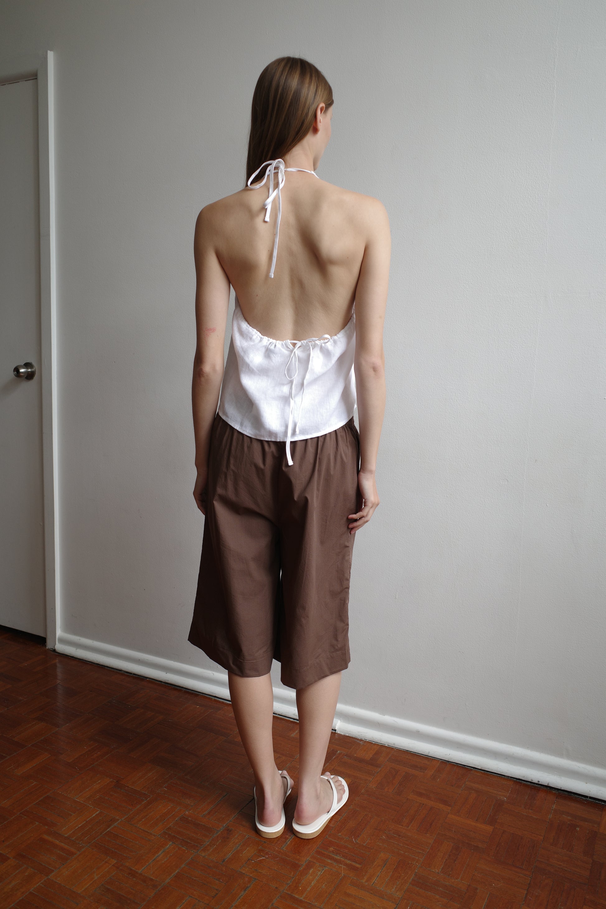 A person stands indoors on a parquet floor, facing a white wall and wearing the Long Lounger - Carob Check from Deiji Studios, styled with a white halter-top in organic cotton and white sandals.
