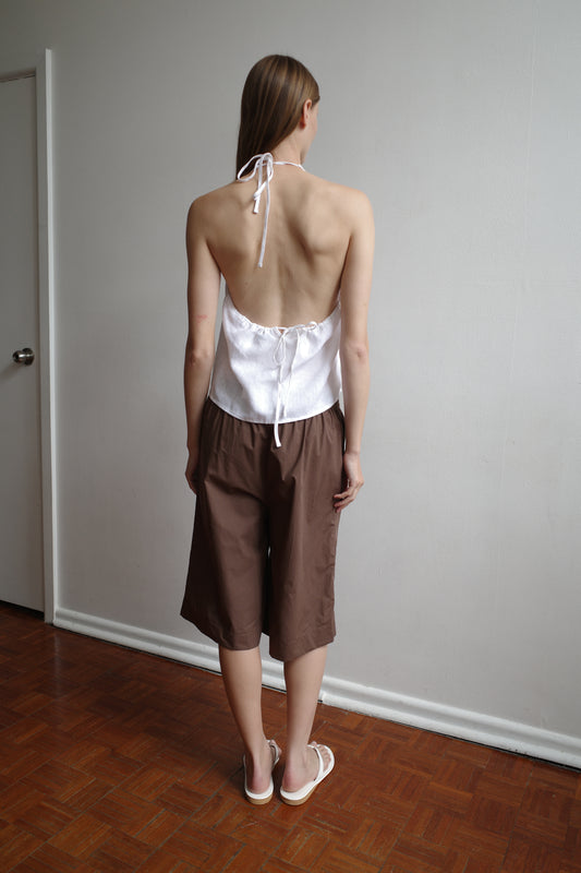 A person stands indoors on a parquet floor, facing a white wall and wearing the Long Lounger - Carob Check from Deiji Studios, styled with a white halter-top in organic cotton and white sandals.