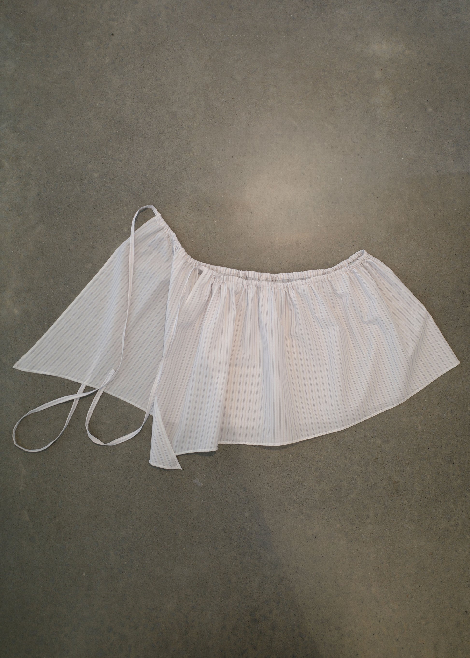 The Napkin Belt - Pacific Stripe, featuring white and light blue stripes in organic cotton with two long tie straps, is displayed flat on a concrete surface.