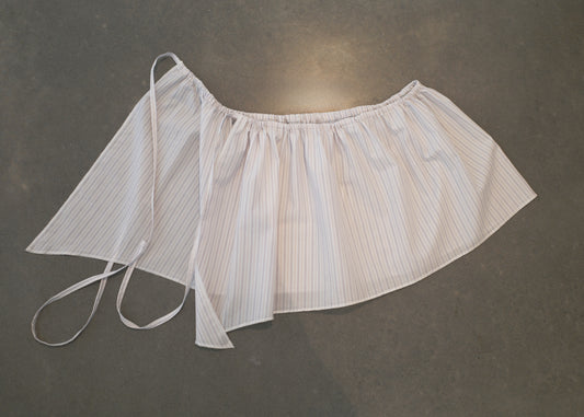 A white and light gray striped, semi-sheer Napkin Belt - Pacific Stripe in organic cotton with gathered fabric and thin ties, laid flat on a smooth, dark surface—crafted with the refined touch of Deiji Studios.