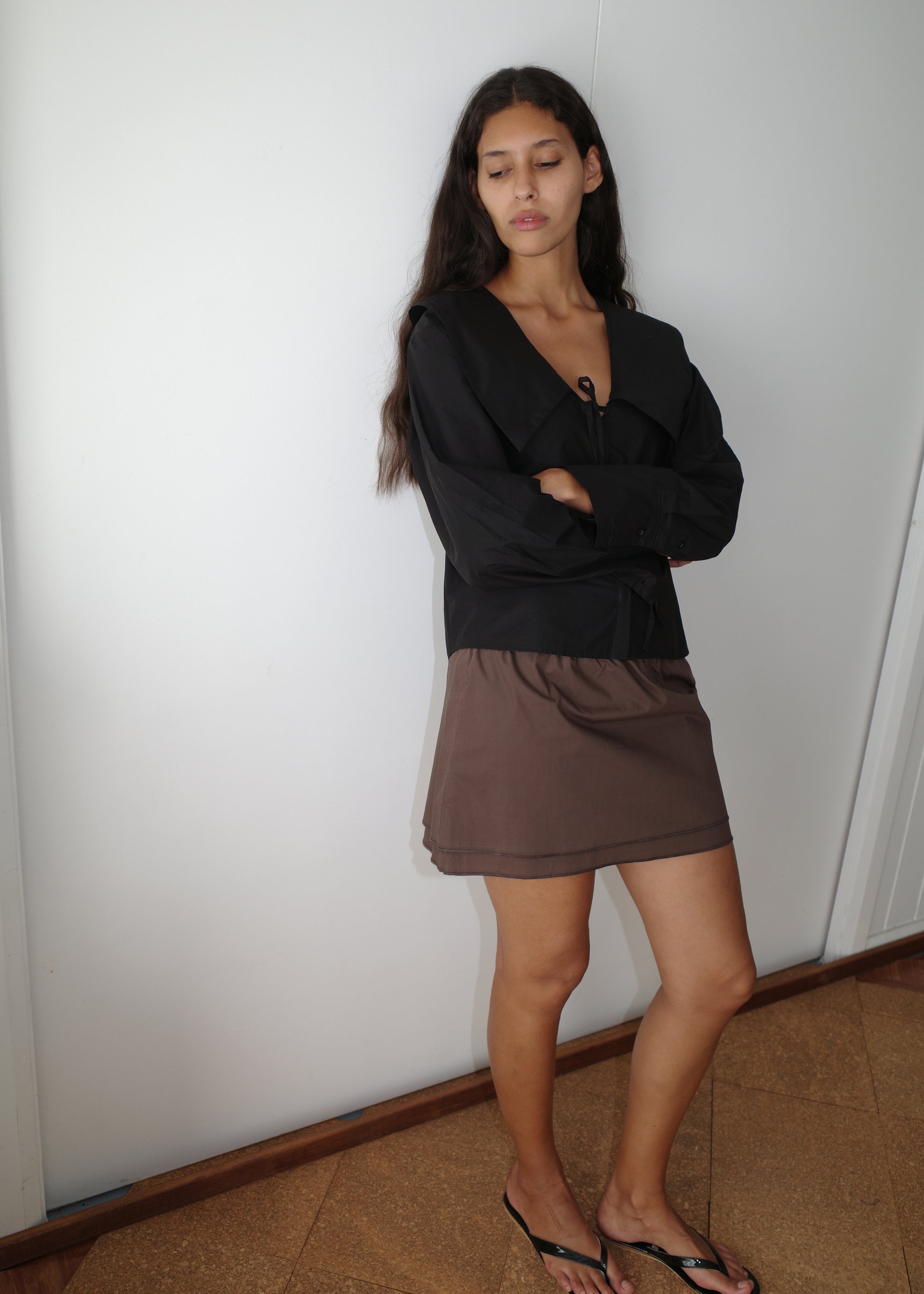 A woman with long dark hair stands against a white wall, arms crossed, wearing the Collared Tie Shirt - Black with a brown skirt and black sandals. She looks down with a neutral expression.