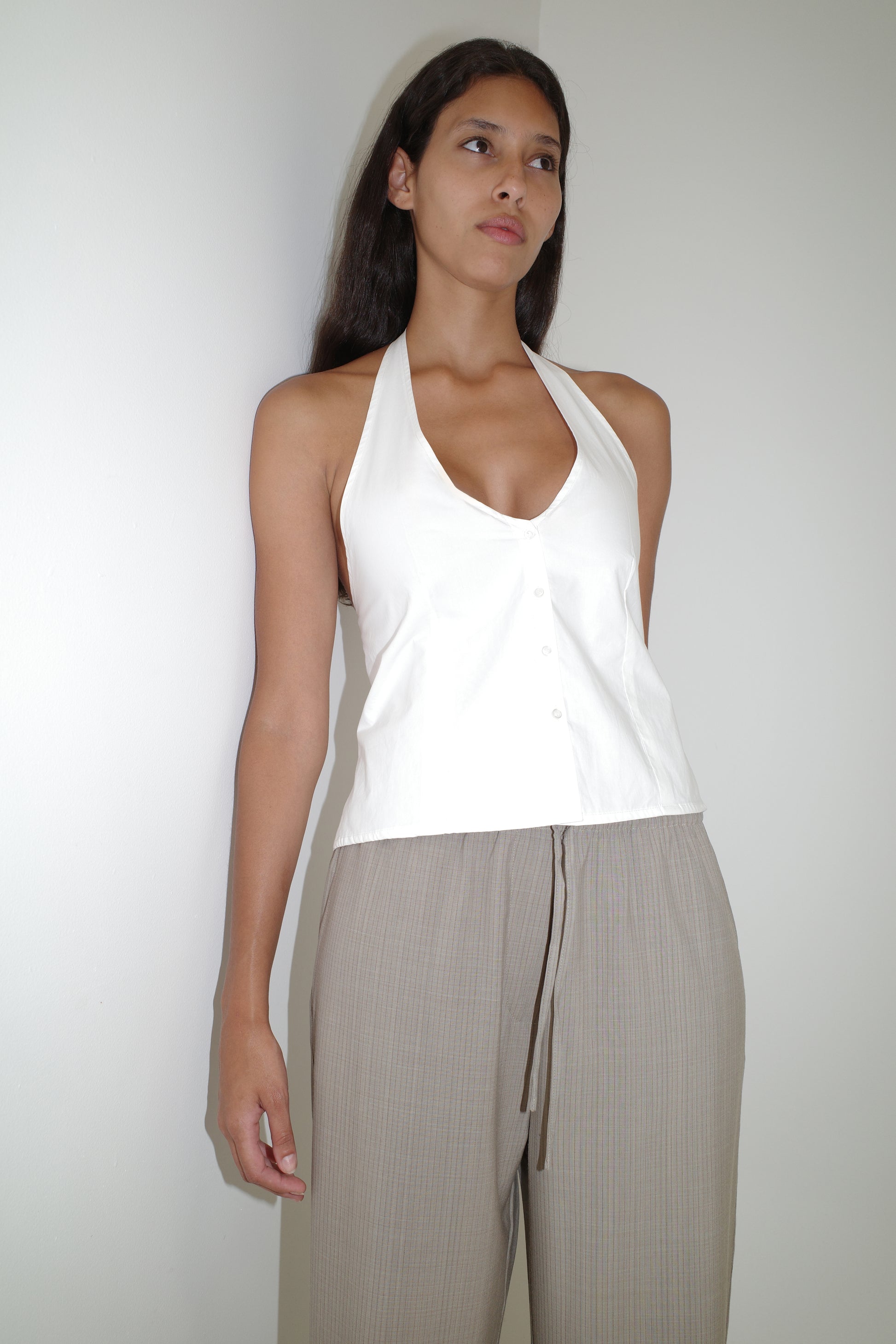 A woman with long dark hair and a neutral expression stands against a white wall, wearing the Low Pocket Pure Wool Pant in Dove Stripe and a white halter-neck top with buttons. The high-waisted pants have a relaxed fit and drawstring detail.