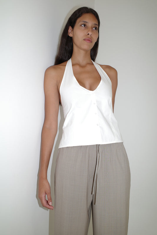 A woman with long dark hair and a neutral expression stands against a white wall, wearing the Low Pocket Pure Wool Pant in Dove Stripe and a white halter-neck top with buttons. The high-waisted pants have a relaxed fit and drawstring detail.