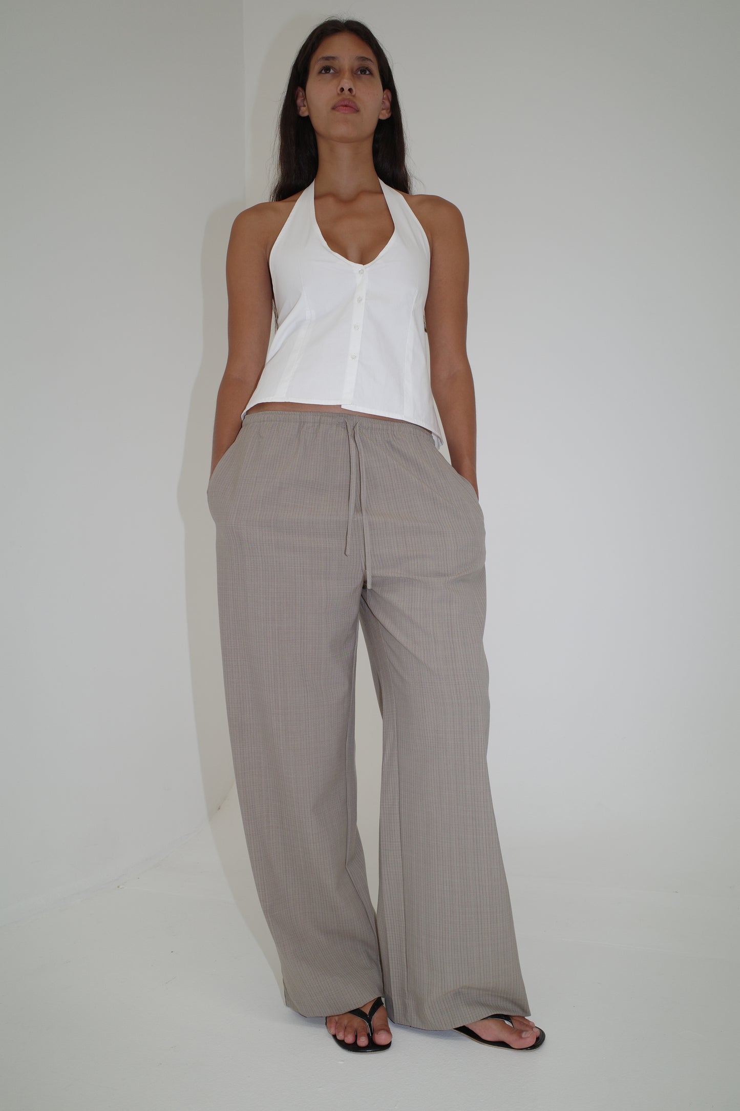 A woman stands against a plain white wall, wearing a sleeveless halter top, black sandals, and the Low Pocket Pure Wool Pant in Dove Stripe. She looks confidently at the camera with her hands in her pockets.
