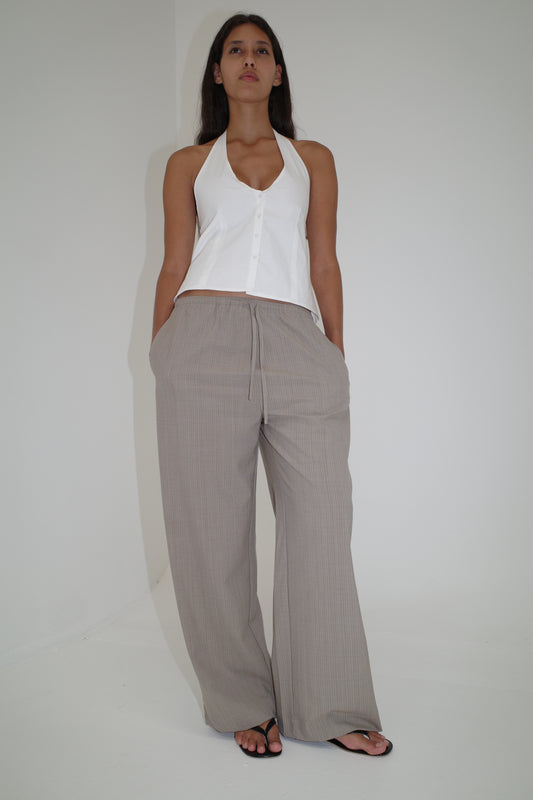 A woman stands against a plain white wall, wearing a sleeveless halter top, black sandals, and the Low Pocket Pure Wool Pant in Dove Stripe. She looks confidently at the camera with her hands in her pockets.