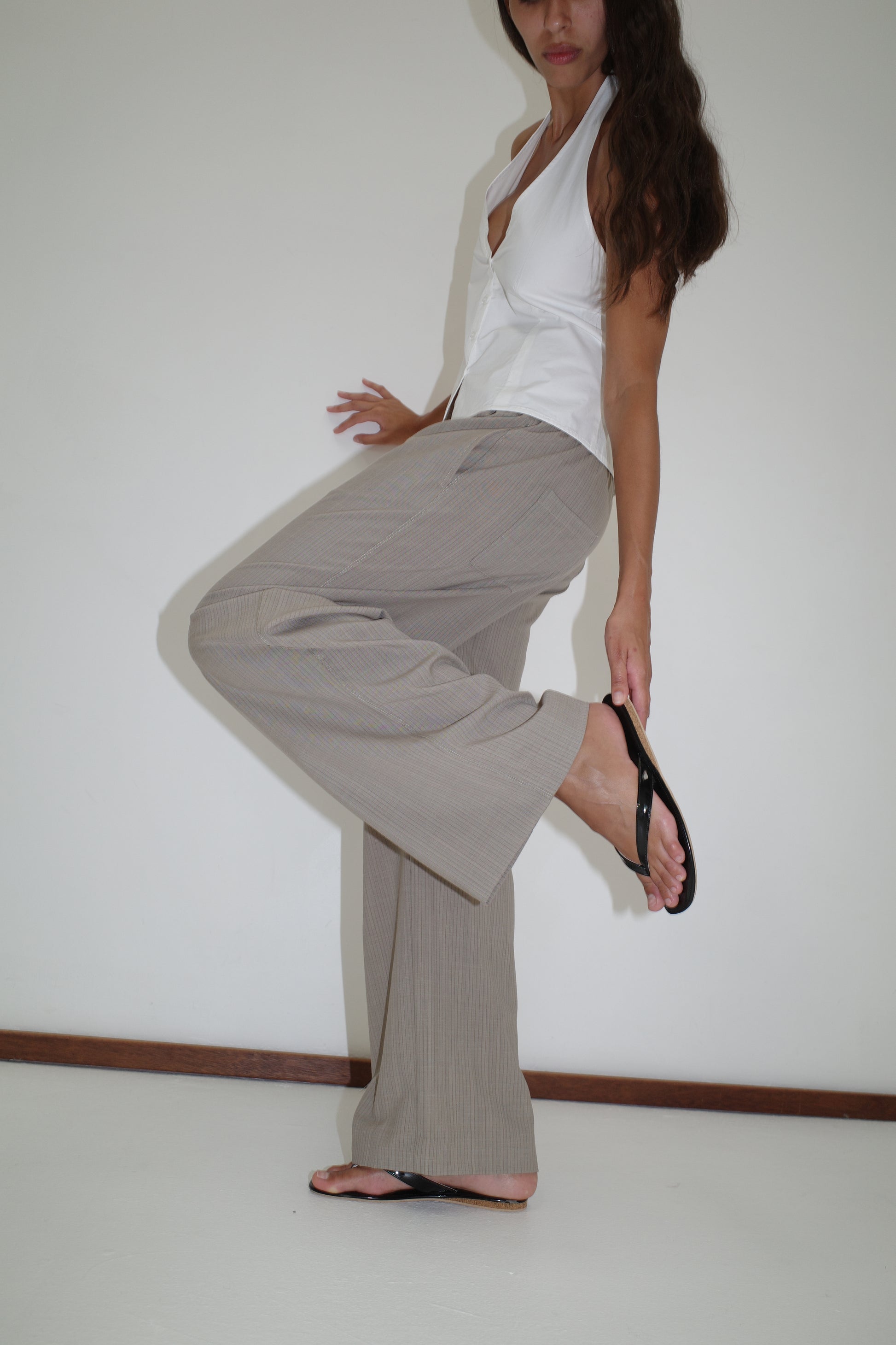 A person wears the Low Pocket Pure Wool Pant in Dove Stripe with a white sleeveless top and black sandals, standing on one leg against a plain white wall and holding their raised foot. Their face is partially out of frame.