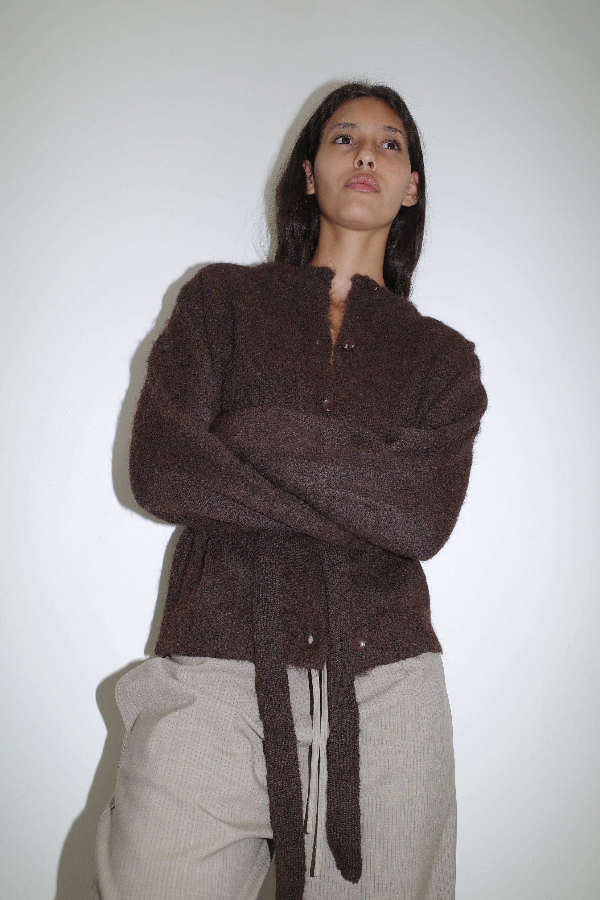 A woman with long dark hair stands against a white wall, arms crossed. She wears the Deiji Studios Belted Cardi - Brown with hanging ties and light beige pants, gazing slightly upward with a serious expression.