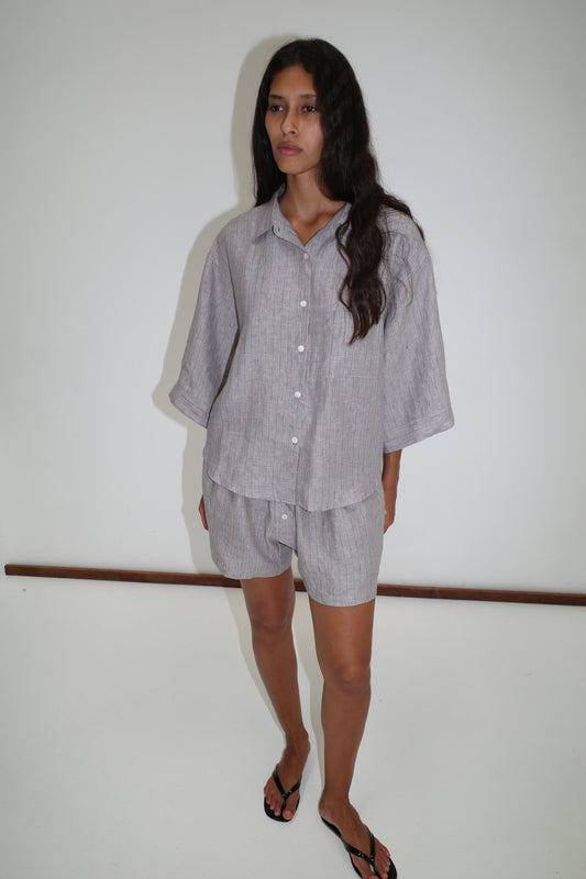 A person with long dark hair wears the 03 Set in Haze Blue, a loose organic French linen button-up shirt and matching boxer shorts, standing barefoot on a white floor against a plain white wall.