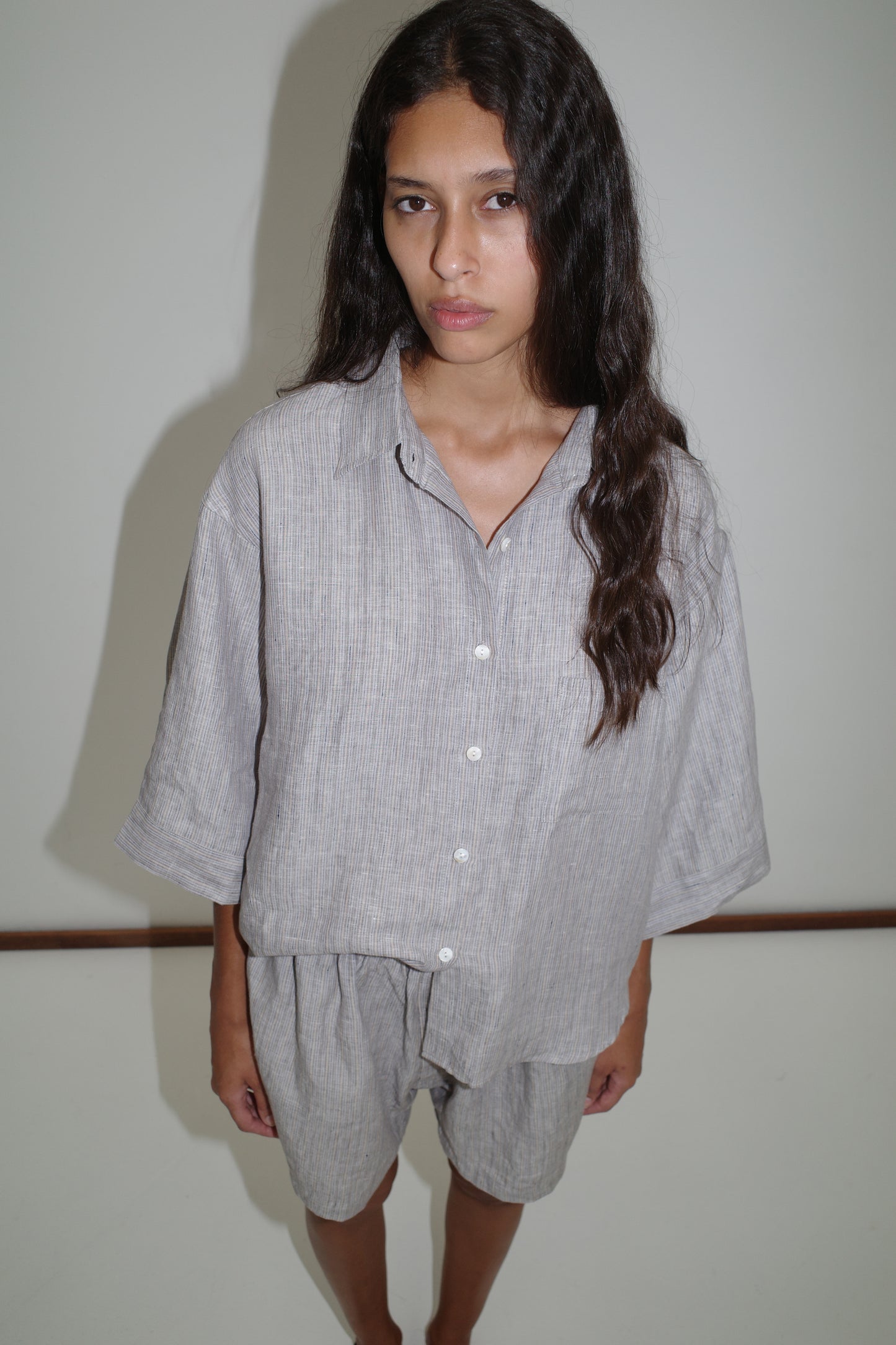 A woman with long dark hair stands against a white wall, wearing the 03 Set - Haze Blue: a loose-fitting organic French linen shirt and matching boxer shorts, looking at the camera with a neutral expression.