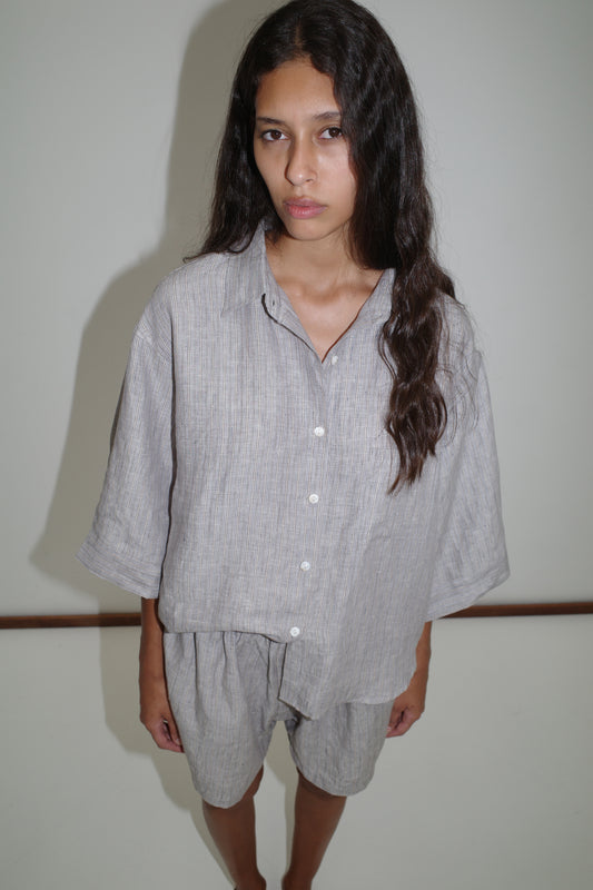 A woman with long dark hair stands against a white wall, wearing the 03 Set - Haze Blue: a loose-fitting organic French linen shirt and matching boxer shorts, looking at the camera with a neutral expression.