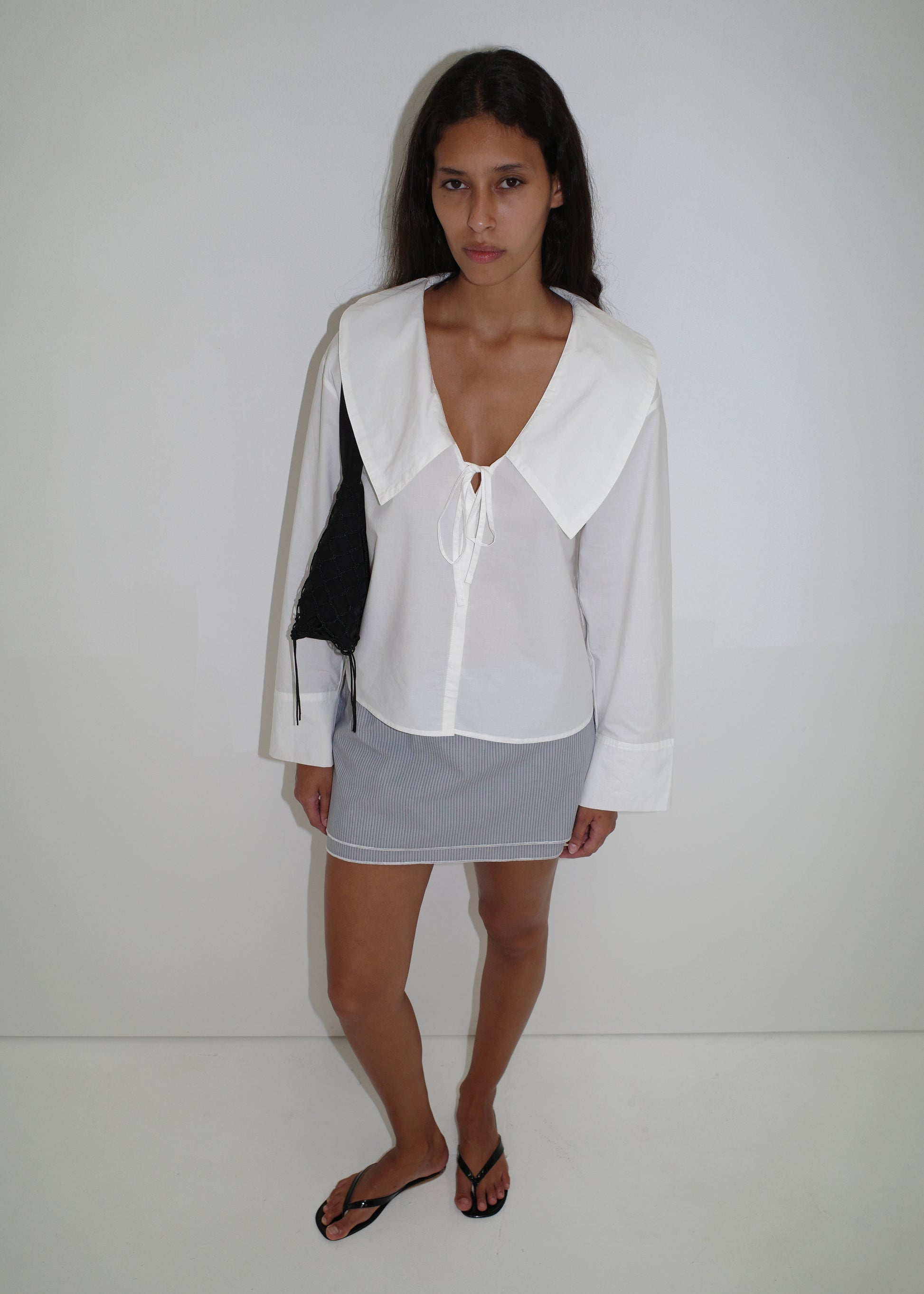 A woman with long dark hair and a neutral expression stands against a white background, wearing the Collared Tie Shirt - White (oversized, organic cotton, exaggerated collar), a light gray mini skirt, black flip-flops, and a black shoulder bag.