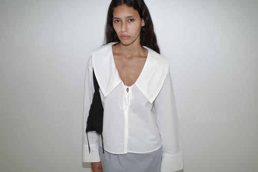 A woman with long dark hair stands against a white wall, wearing the Collared Tie Shirt - White, a light gray skirt, and carrying a black shoulder bag. She looks directly at the camera with a neutral expression.