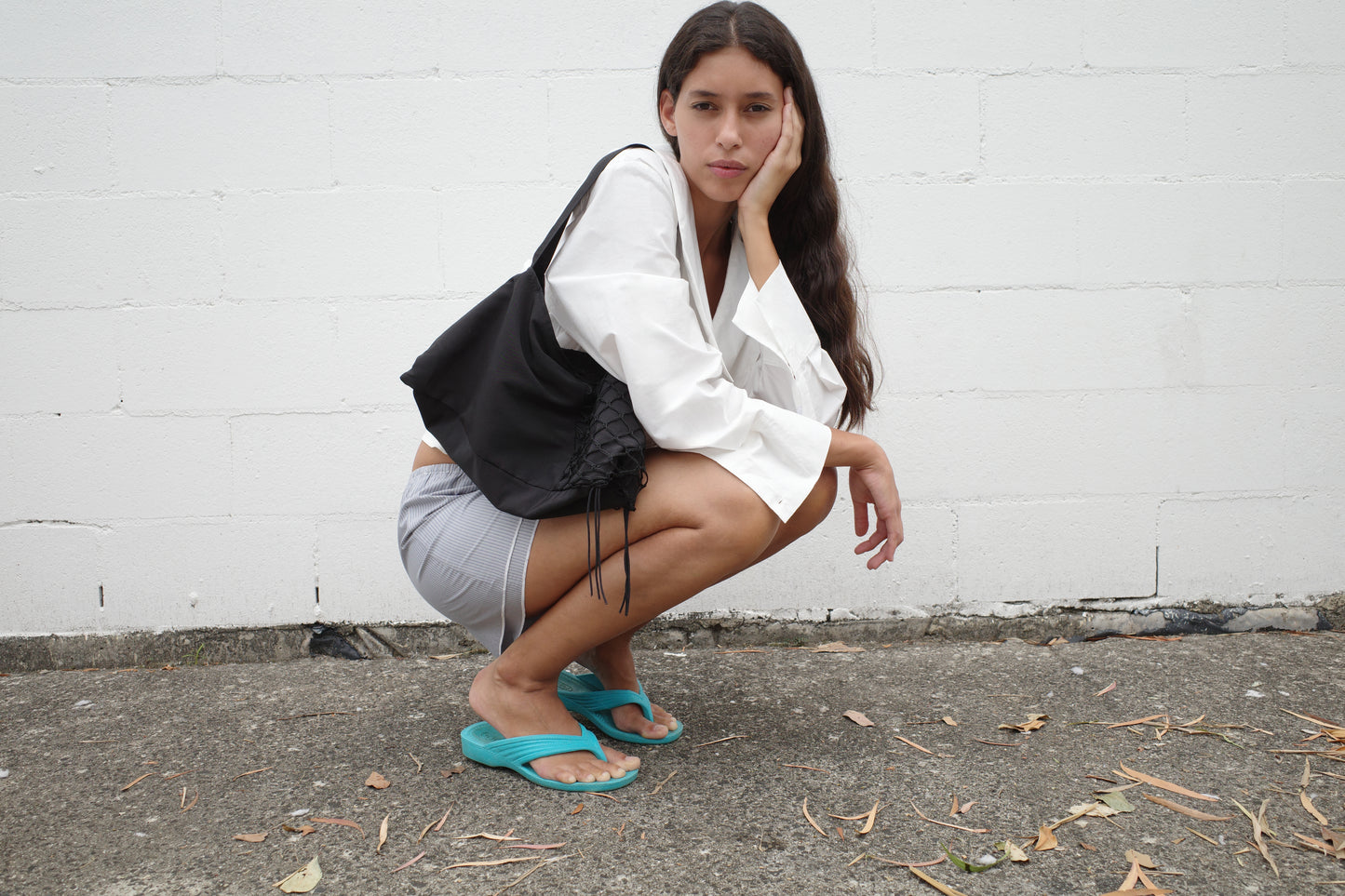 A woman crouches by a white brick wall, wearing a white Collared Tie Shirt made from organic cotton, gray shorts, teal flip-flops, and holding a black drawstring bag while resting her cheek on her hand.