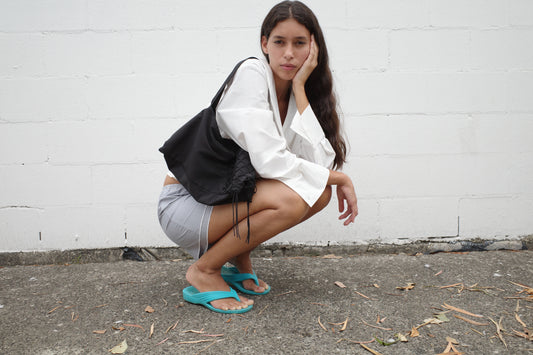 A person with long hair squats outdoors against a white brick wall, wearing the Collared Tie Shirt - White, gray shorts, turquoise flip-flops, and holding a black bag. They rest their face on one hand while looking at the camera.