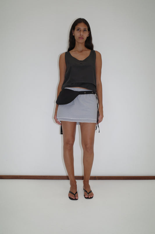 A woman with long dark hair and a neutral expression stands against a white wall, wearing the Layered Top - Black Stripe, a short light blue skirt with white trim, black waist bag, and sandals for a relaxed, comfortable look.