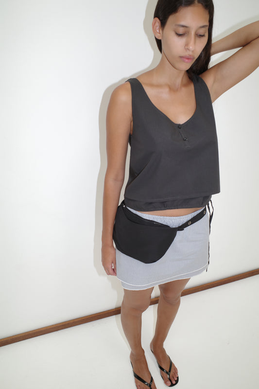 A woman stands against a white wall, wearing the Layered Top - Black Stripe from Deiji Studios, a light mini skirt, black sandals, and a black waist bag. One arm is raised as she looks down with a neutral expression.