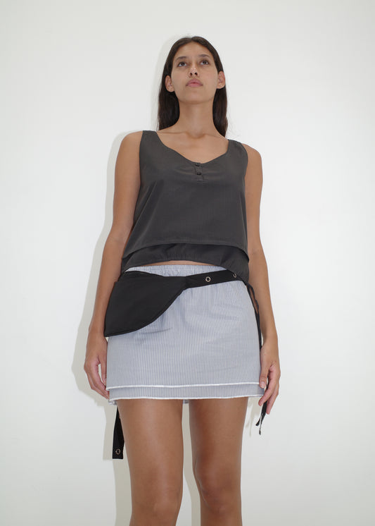 A woman with long dark hair wears the Layered Top - Black Stripe against a white background, paired with a light striped mini skirt and a black belt bag, and shows a neutral expression.