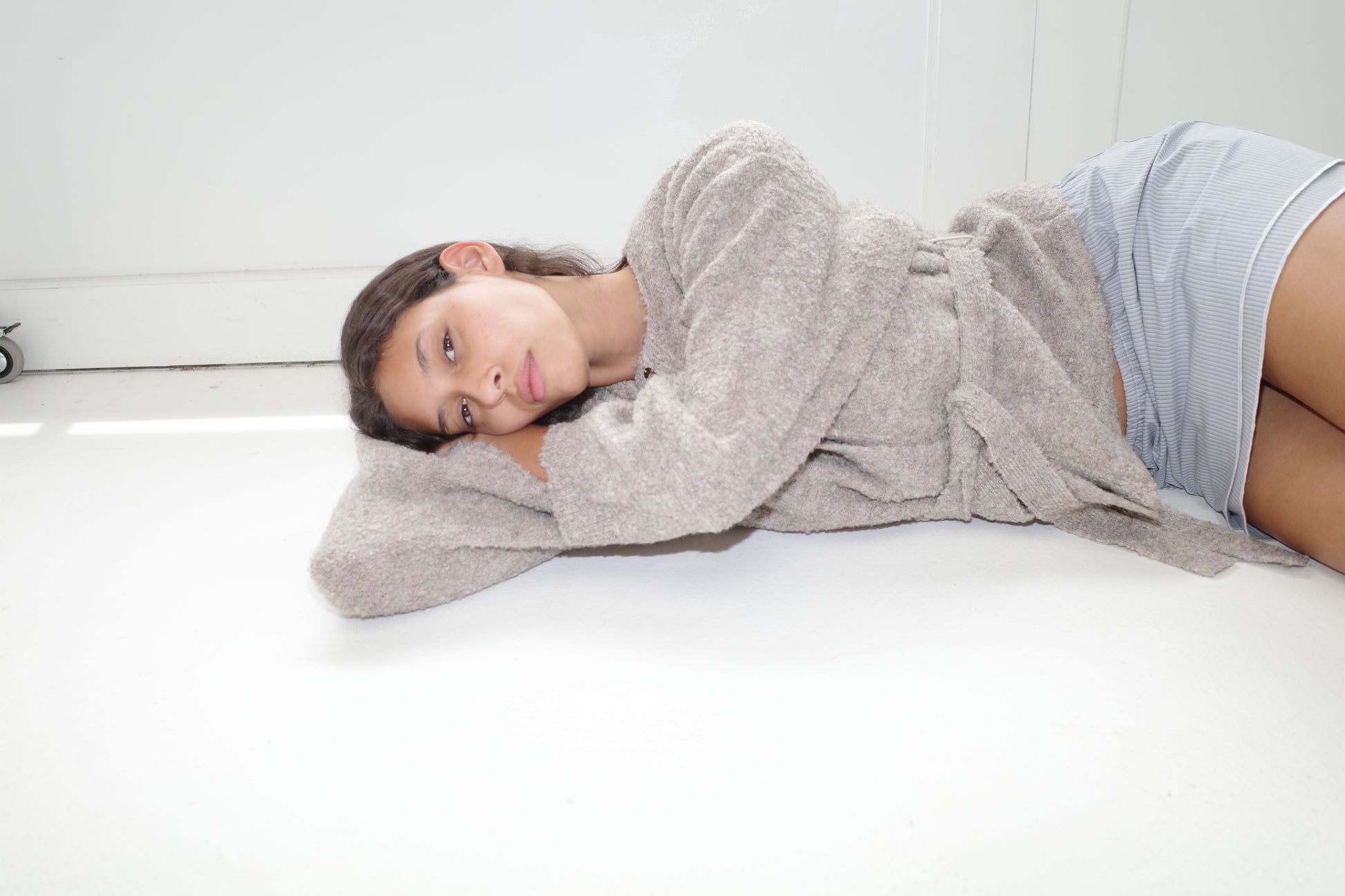 A young woman in Deiji Studios' Pod Knit - Cloud Grey robe and striped shorts lies on her side on a white floor, resting her head on her arm and looking toward the camera, embodying sustainable comfort.